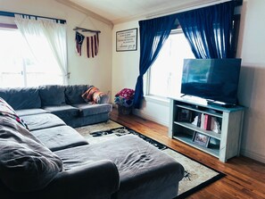 A living room with a grey sofa, a flat-screen TV on a wooden stand, and a window with blue curtains.