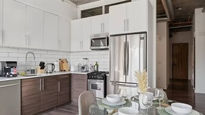 Cozinha moderna com armários brancos, eletrodomésticos de aço inoxidável e área de jantar com mesa de vidro para quatro pessoas.