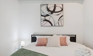 Een net opgemaakt bed met hoofdbord, een aan de muur bevestigd abstract schilderij en een bedlamp.