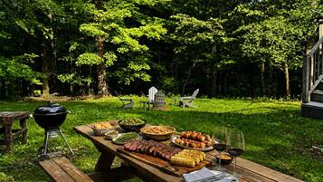 Un espace extérieur aménagé pour les repas, avec une table de pique-nique dressée, entouré d’une végétation luxuriante et d’arbres.