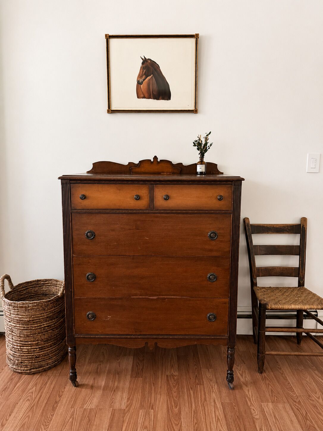 Een houten dressoir met laden en een spiegel, een rieten mand, en een houten stoel.