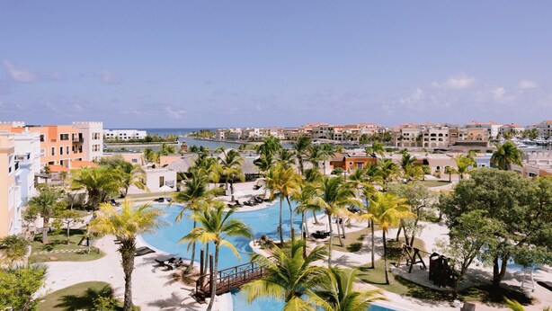 A resort with a swimming pool surrounded by palm trees and a view of the ocean. - Marina View 2BR Apartment  Cap Cana (Punta Cana)