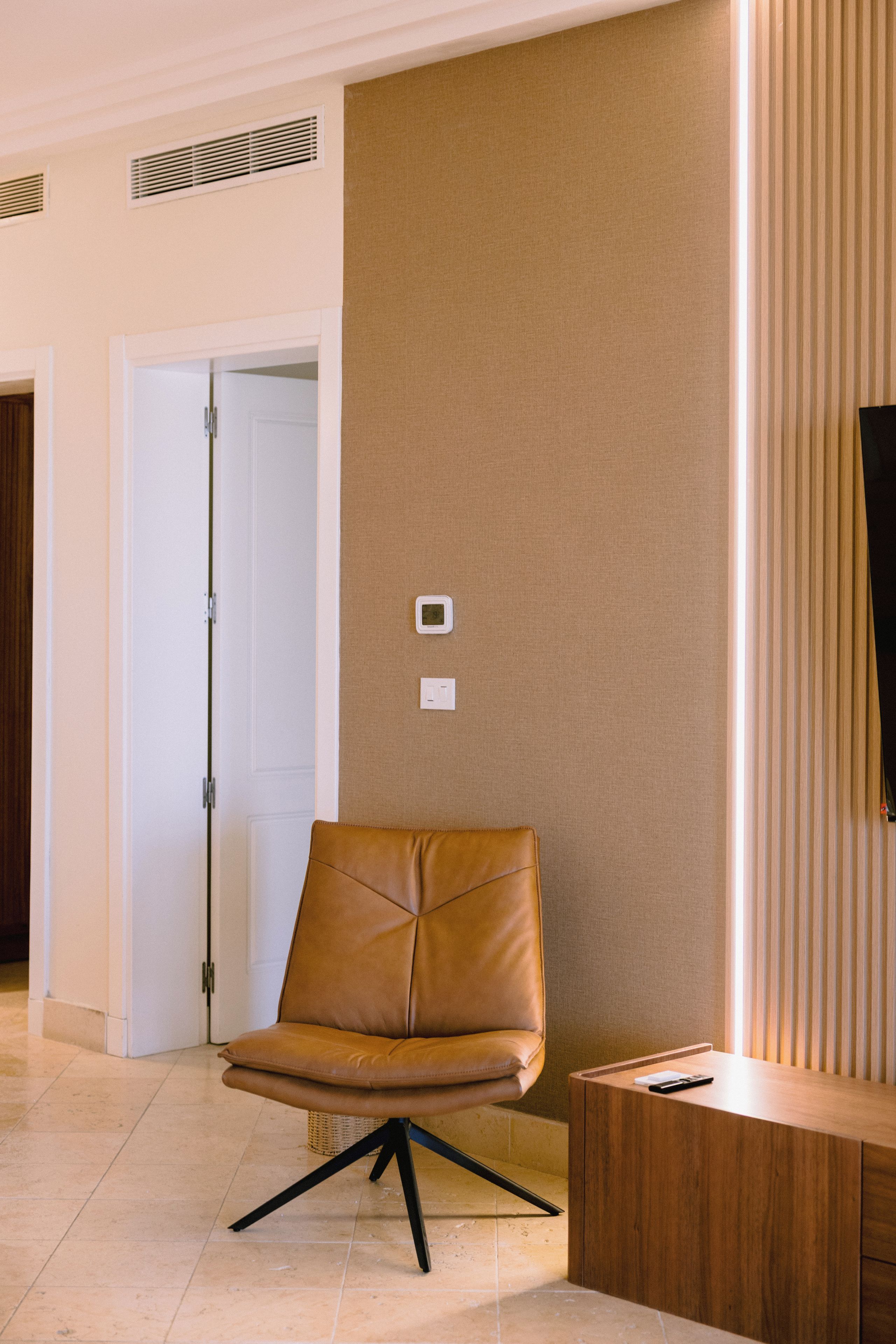 Une chaise moderne avec un siège en cuir et des pieds en métal noir, placée dans un couloir où se trouvent une console en bois et un téléviseur posé sur un meuble.