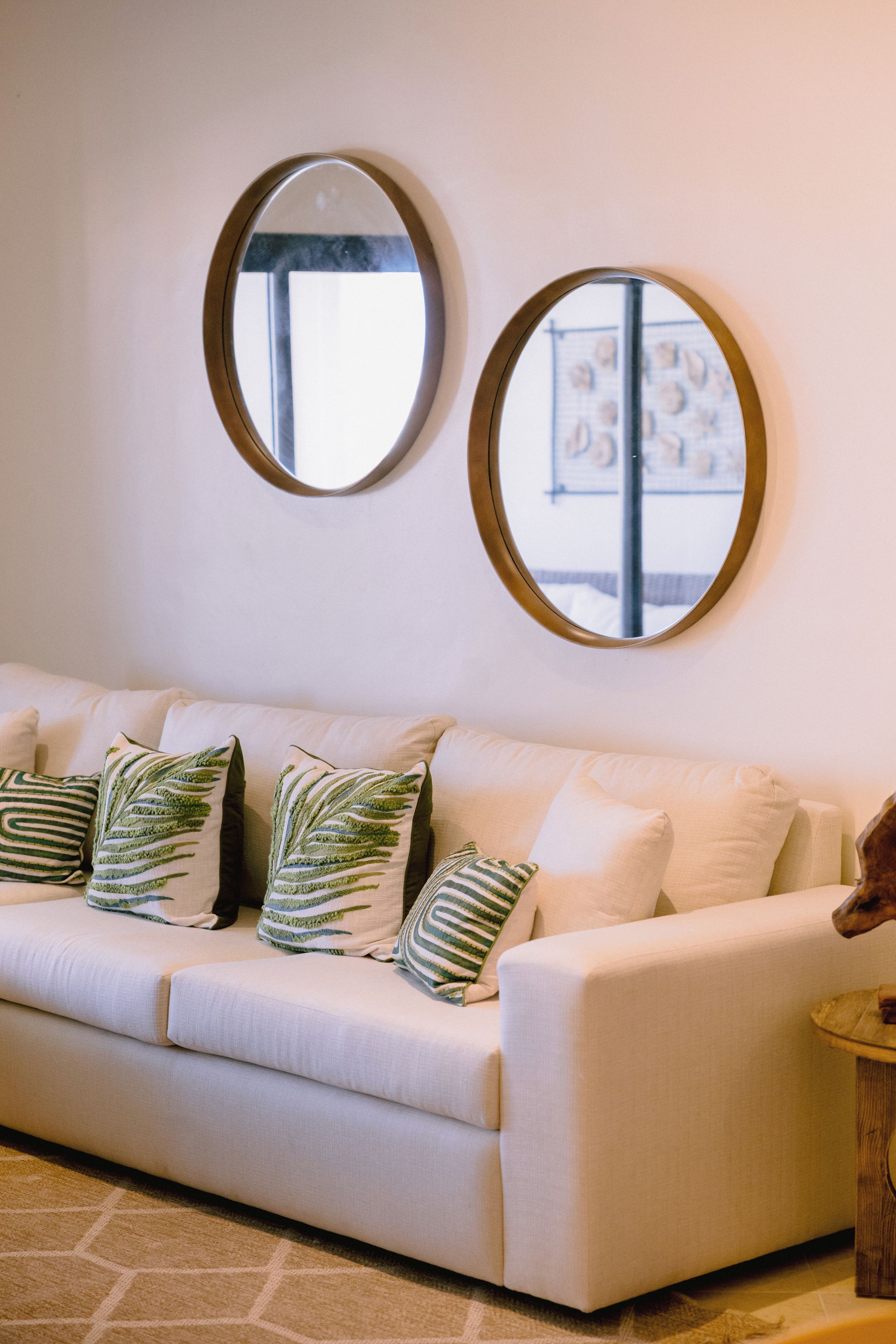 A white sofa with green and white patterned pillows, two round mirrors on the wall, and a patterned rug on the floor.