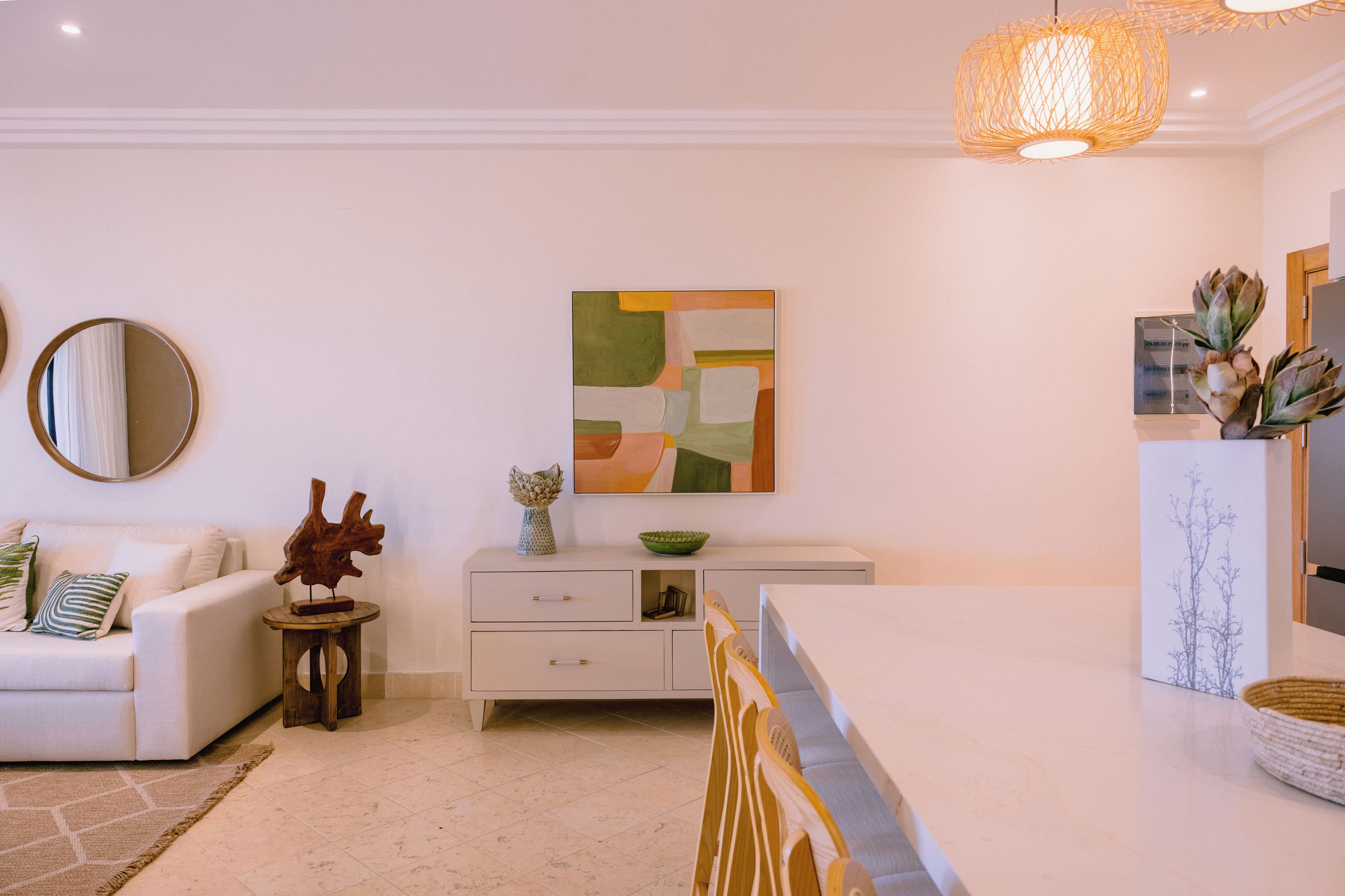 Un salon moderne avec un canapé blanc, un miroir rond, une table d’appoint en bois, un meuble blanc avec des tiroirs et un tableau abstrait suspendu au mur.