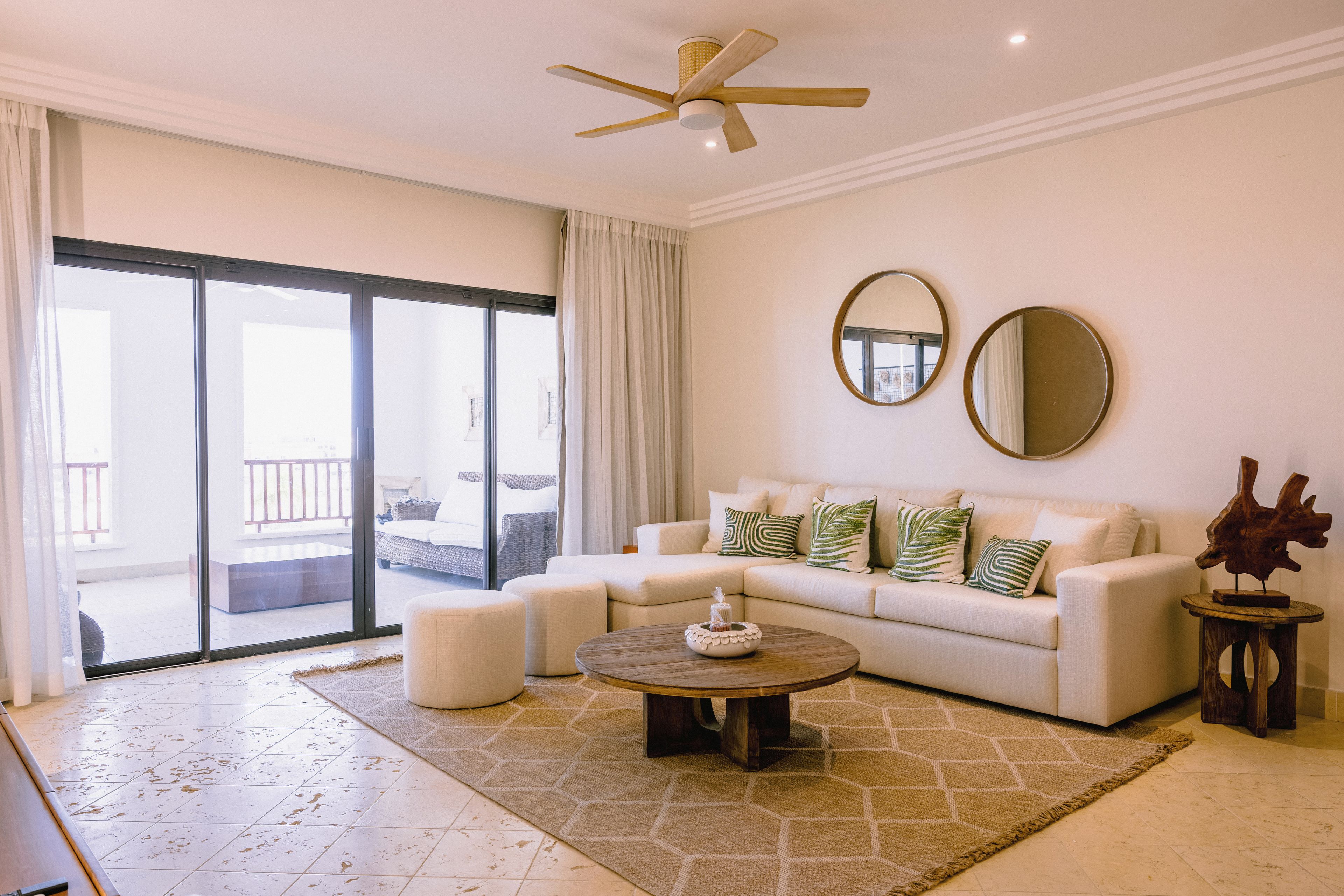 A spacious living room with a large sliding glass door leading to a balcony, a white sofa with patterned cushions, a round wooden coffee table, and two wall-mounted mirrors.