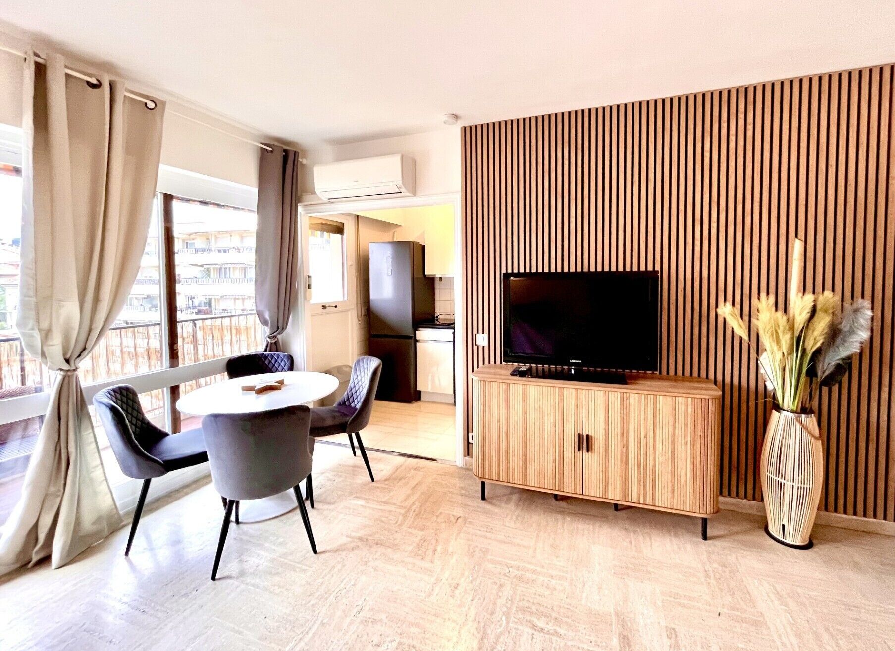 A modern living room with a flat-screen TV, a dining area with two chairs, a wooden cabinet, and a vase with dried plants.