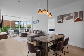 A modern living room with a dining area, large windows, and a view of greenery outside. - Beachside Stay in Kelowna | Pool, Gym & Sauna (Kelowna)