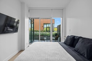 A modern bedroom with a bed, a sofa, a TV, and a sliding glass door leading to an outdoor area with a table and chairs.