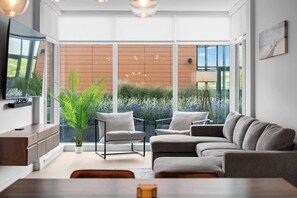 Una sala de estar moderna con un sofá, sillones, un televisor y un ventanal grande con vista a un jardín.
