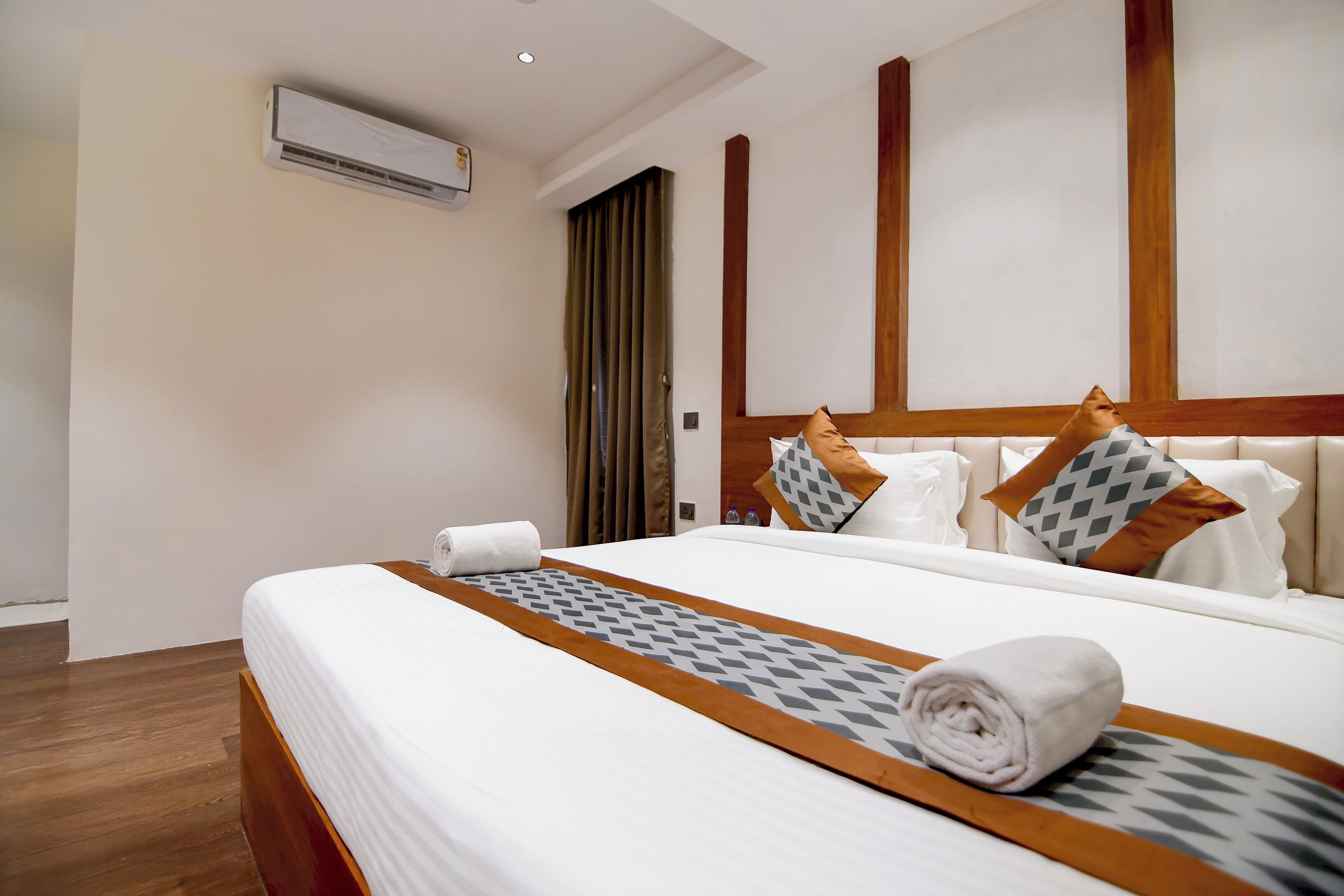 A hotel room with a bed, a wooden headboard, a window with curtains, and an air conditioning unit on the wall.