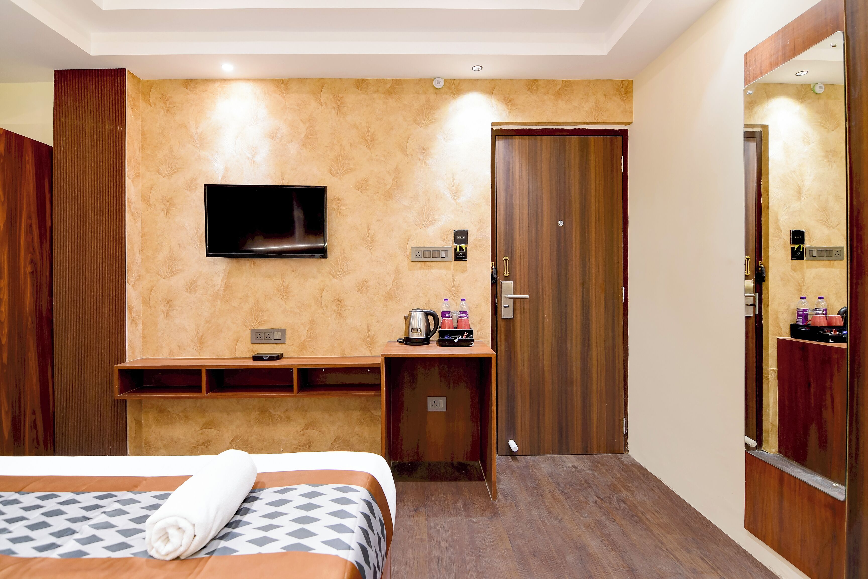 Habitación de hotel con cama, televisor montado en la pared, escritorio con hervidor y botellas, y puerta de madera.