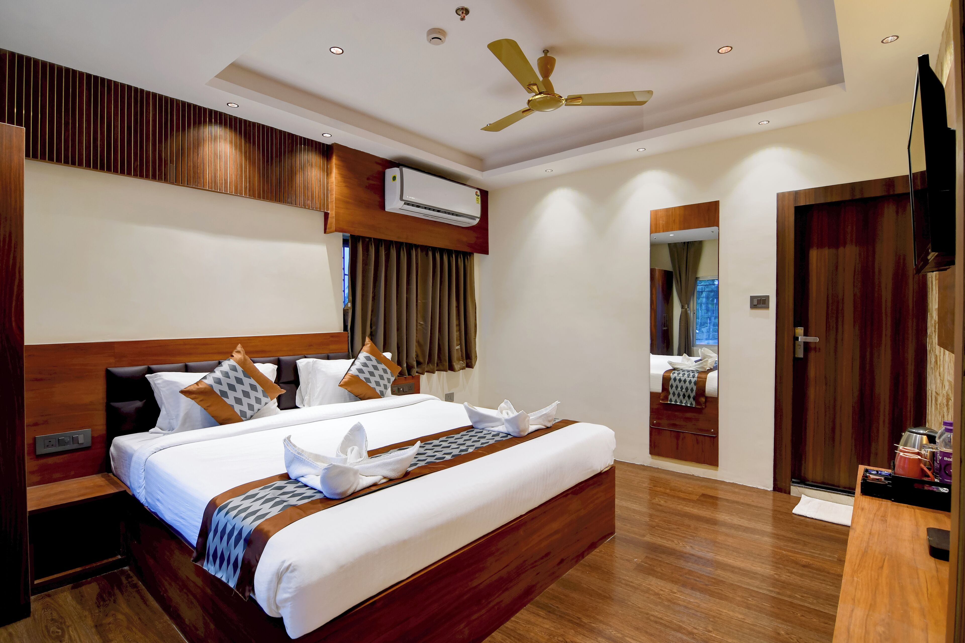 A hotel room with a large bed, a ceiling fan, an air conditioning unit, a mirror, and a wooden floor.