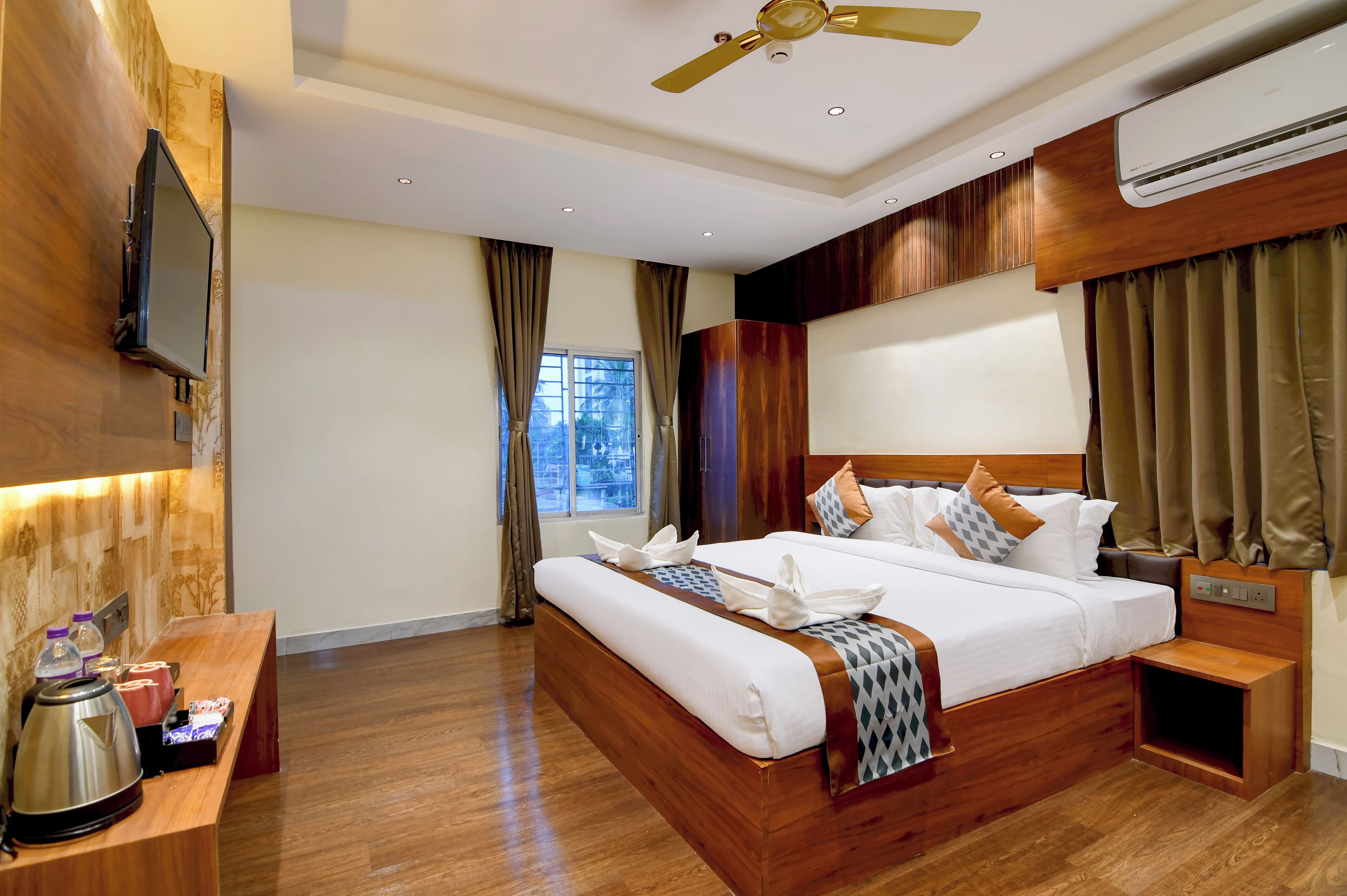 Ein Hotelzimmer mit einem großen Bett, einem flachbildfernseher an der Wand, einem Deckenventilator und einem Fenster mit Vorhängen.