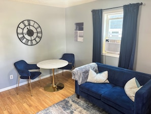 A living room with a blue sofa, two blue chairs, a round table, a wall clock, and a window with curtains. - Perfect 2-bedroom house with AC in welcoming East Grand Forks (East Grand Forks)