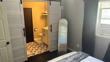 Un dormitorio con cama, puerta corredera para armario, baño con inodoro y lavamanos, y ventana con cortinas.
