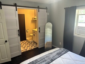 A bedroom with a bed, a sliding closet door, a bathroom with a toilet and sink, and a window with curtains.