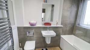 A modern bathroom with a white sink, toilet, and bathtub. There is a scale on the floor, a towel rack, and a mirror above the sink. - 4 Goldcrest Close (Daventry)