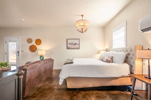 Een slaapkamer met een groot bed, een nachtkastje, een dressoir, een kroonluchter en een raam met jaloezieën.