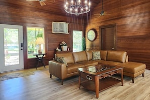A living room with a brown leather sectional sofa, a glass coffee table, wooden walls, a clock, and a door leading outside. - Clear Lake Waterfront • Between NASA & Kemah (Seabrook)