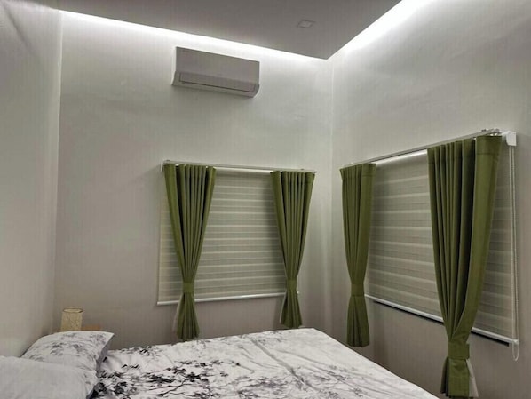 A bedroom with a bed, two windows with curtains, an air conditioning unit, and a ceiling light.