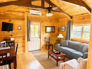 A cozy cabin interior with a wooden ceiling, a ceiling fan, a television mounted on the wall, a sofa, a coffee table, and a dining area with chairs and a table.