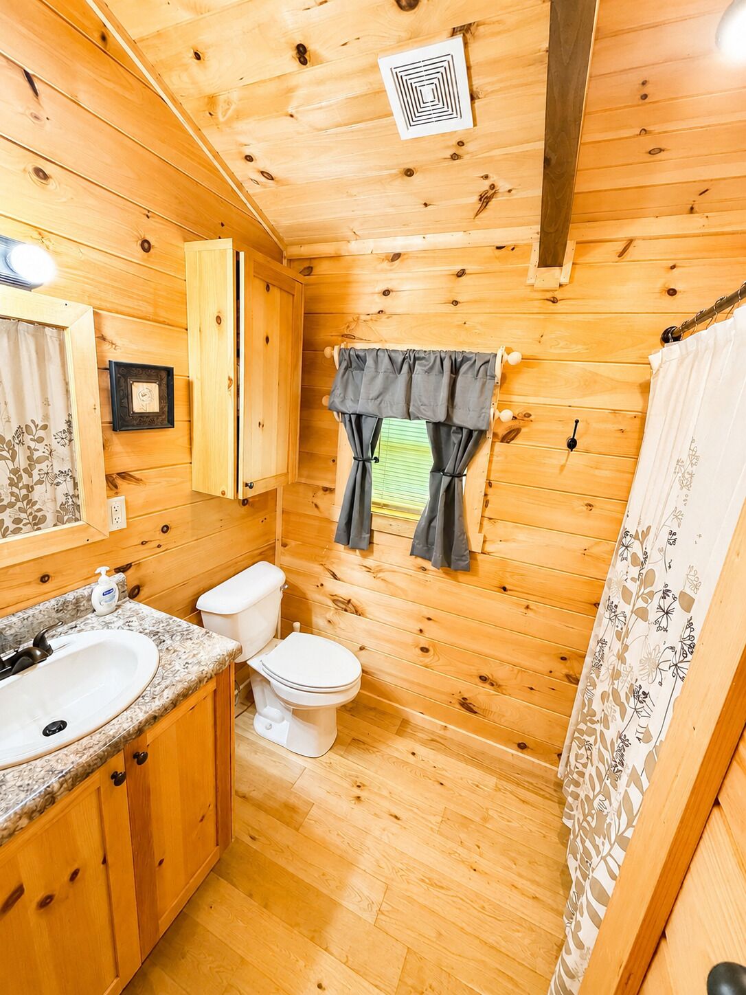 A wooden cabin bathroom with a sink, toilet, and window with curtains.