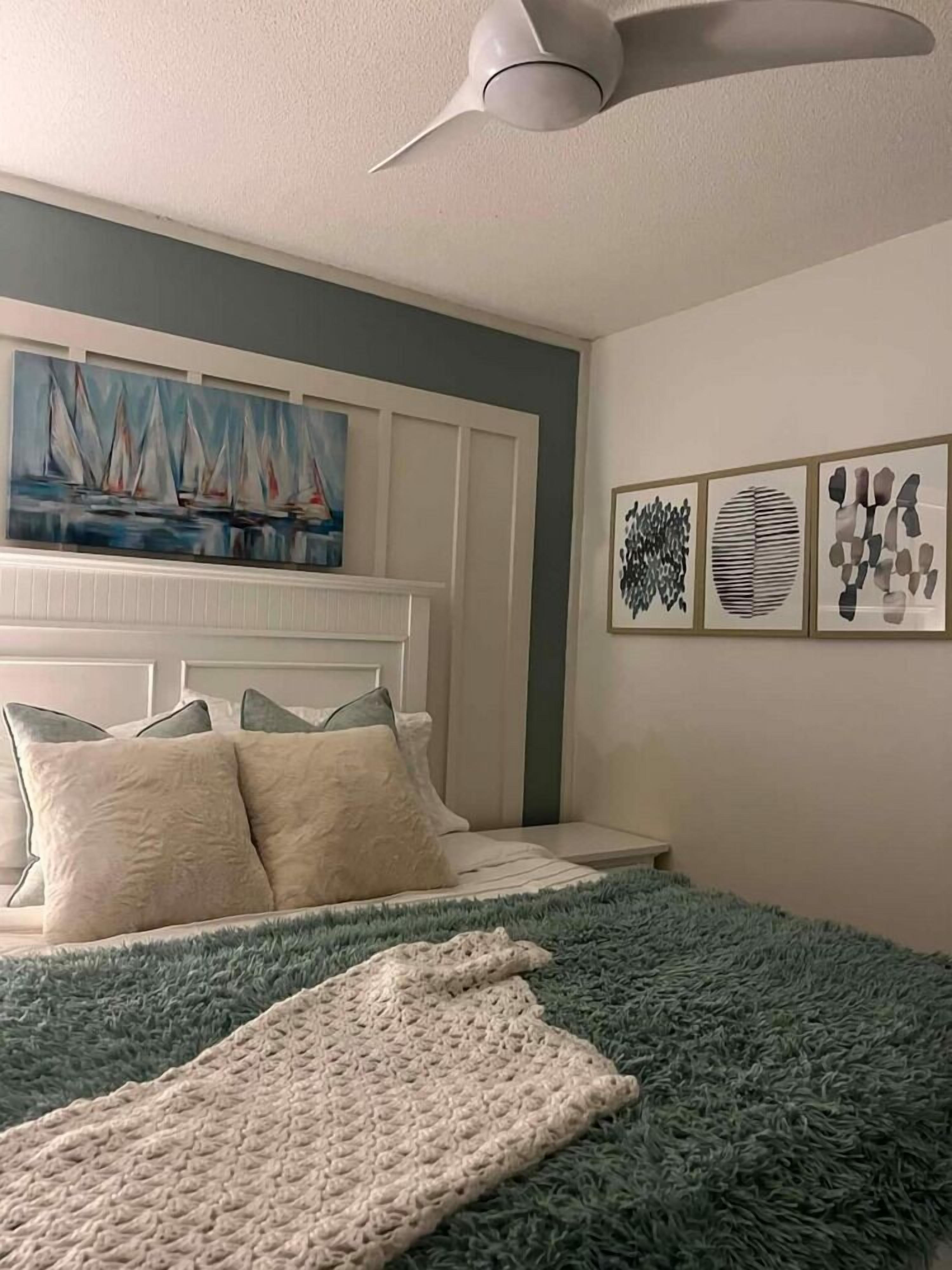 Quarto com cama, ventilador de teto, pintura de veleiros e arte abstrata na parede.