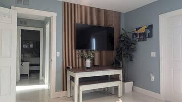 Una habitación con un televisor montado en la pared, una mesita consola de madera, una planta en maceta y una puerta que da a otra habitación.