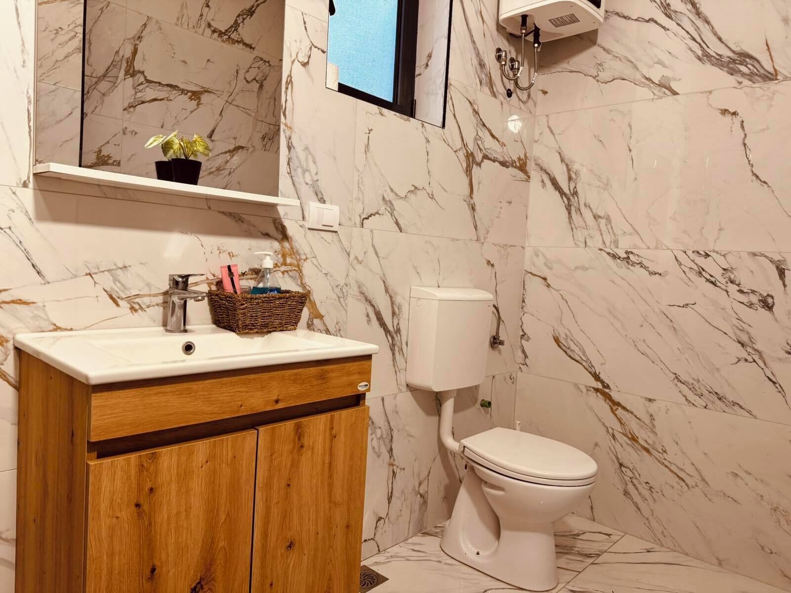 A bathroom with a marble wall, a wooden vanity, a white toilet, and a small shelf with toiletries.
