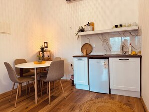 Een kleine keuken met een eettafel en stoelen, een kleine koelkast en een aanrecht met verschillende spullen.