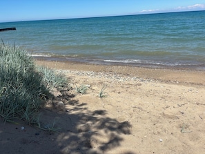 Ein sandiger Strand mit sanften Wellen, blauem Himmel und spärlicher Vegetation.