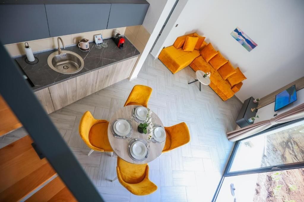 A modern kitchen with a sink, a dining area with a round table and yellow chairs, and a bright orange sofa in a living space.