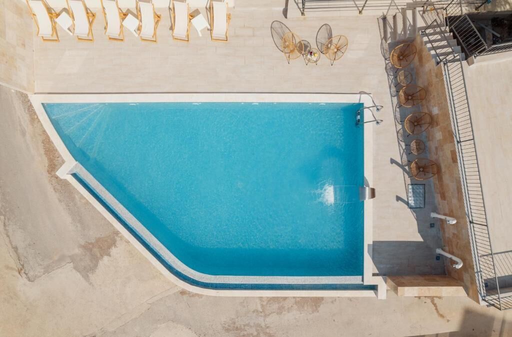 Vista aérea de una piscina con borde curvo, rodeada por una terraza con baldosas y algunas sillas de descanso.