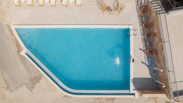 Vista aérea de una piscina con borde curvo, rodeada por una terraza con baldosas y algunas sillas de descanso.