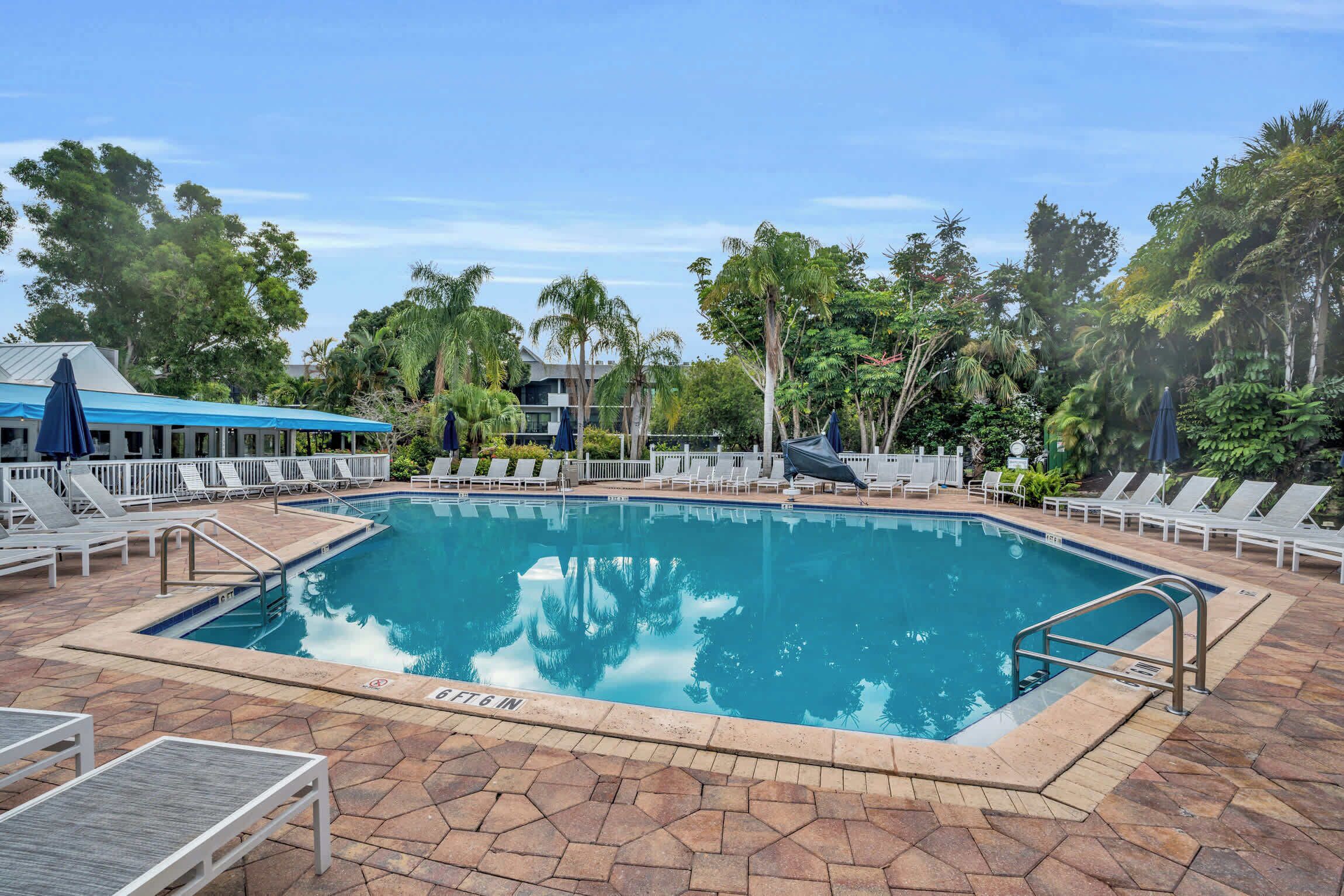 A rectangular swimming pool with a clear blue water surface, surrounded by a tiled deck with lounge chairs and umbrellas.
