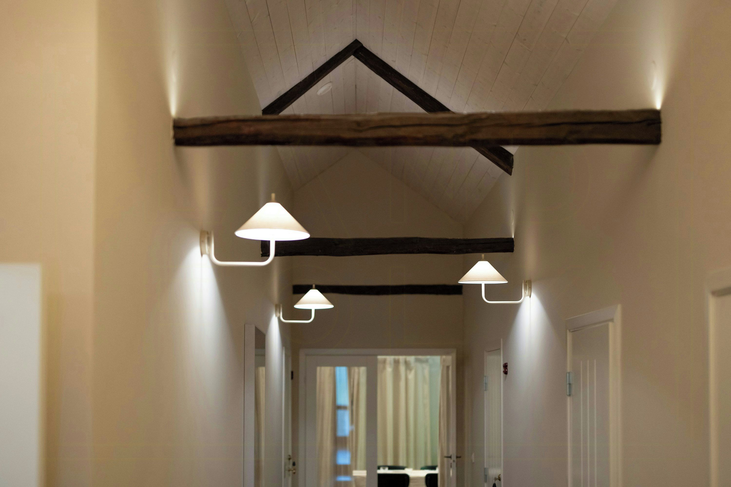 A hallway with exposed wooden beams, white walls, and three wall-mounted lamps.