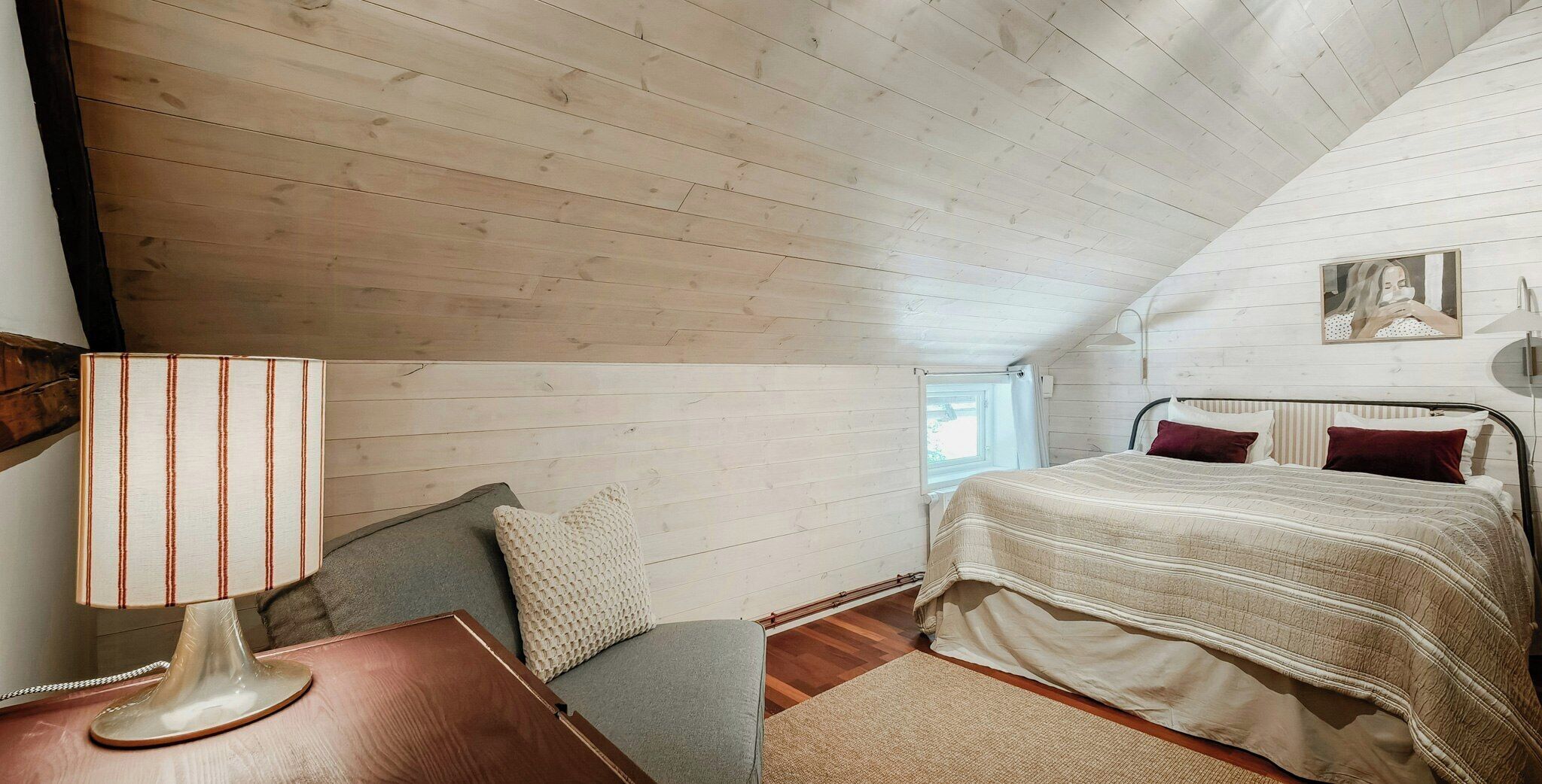 Une chambre avec un plafond en pente, un lit recouvert d’une couvre-lit à rayures, une table de chevet avec une lampe et une fenêtre.