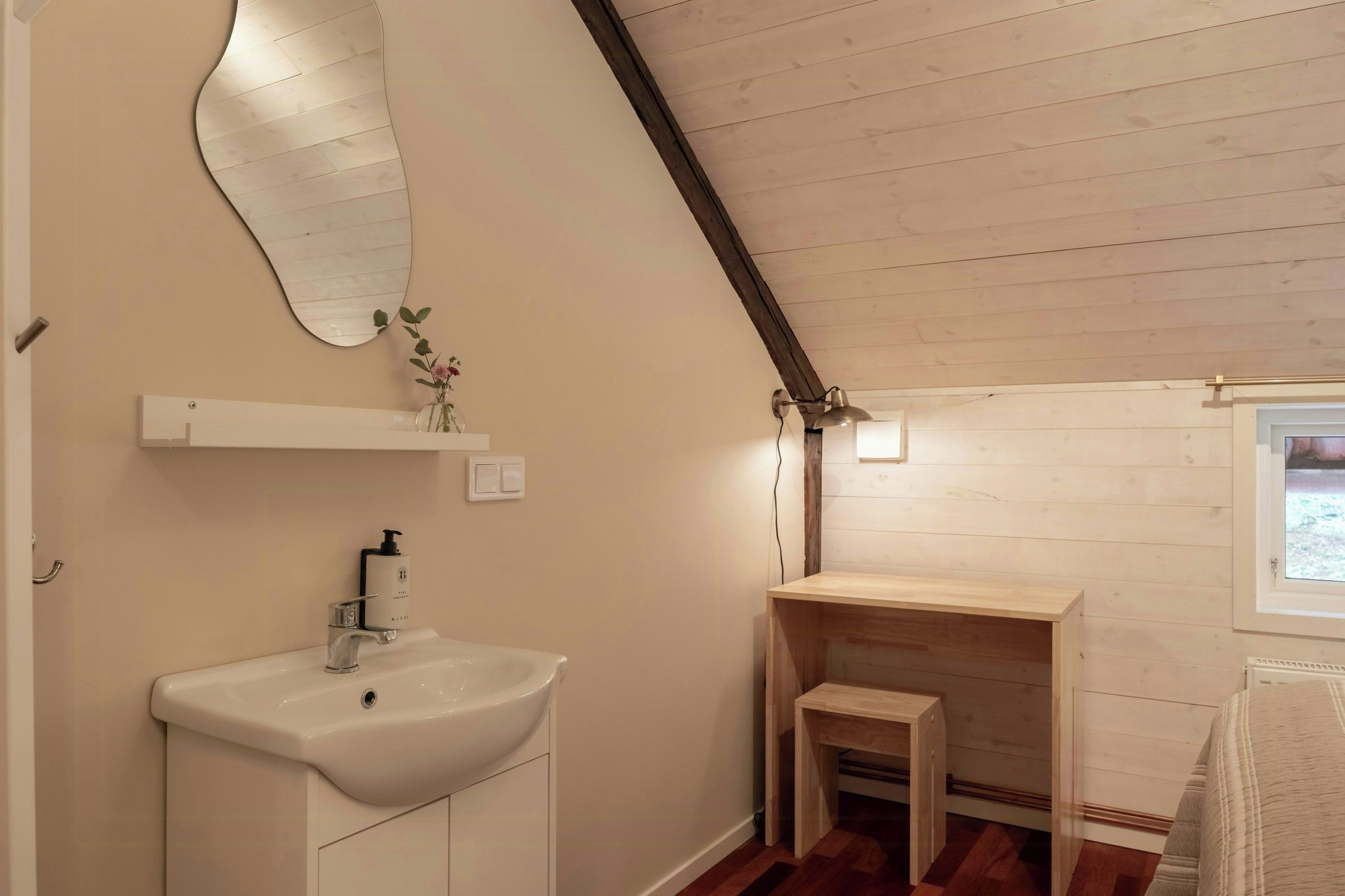 A bathroom with a white sink, a mirror, a shelf with a vase, and a wooden bench.