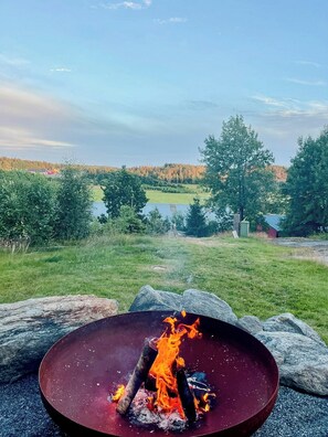 Un feu de camp dans un récipient métallique, entouré de pierres, installé dans un champ herbeux avec des arbres et une vue lointaine sur un lac et des bâtiments.