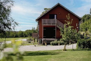 En tvåvånings ladugård i trä med en balkong, omgiven av grönska och en grusuppfart.