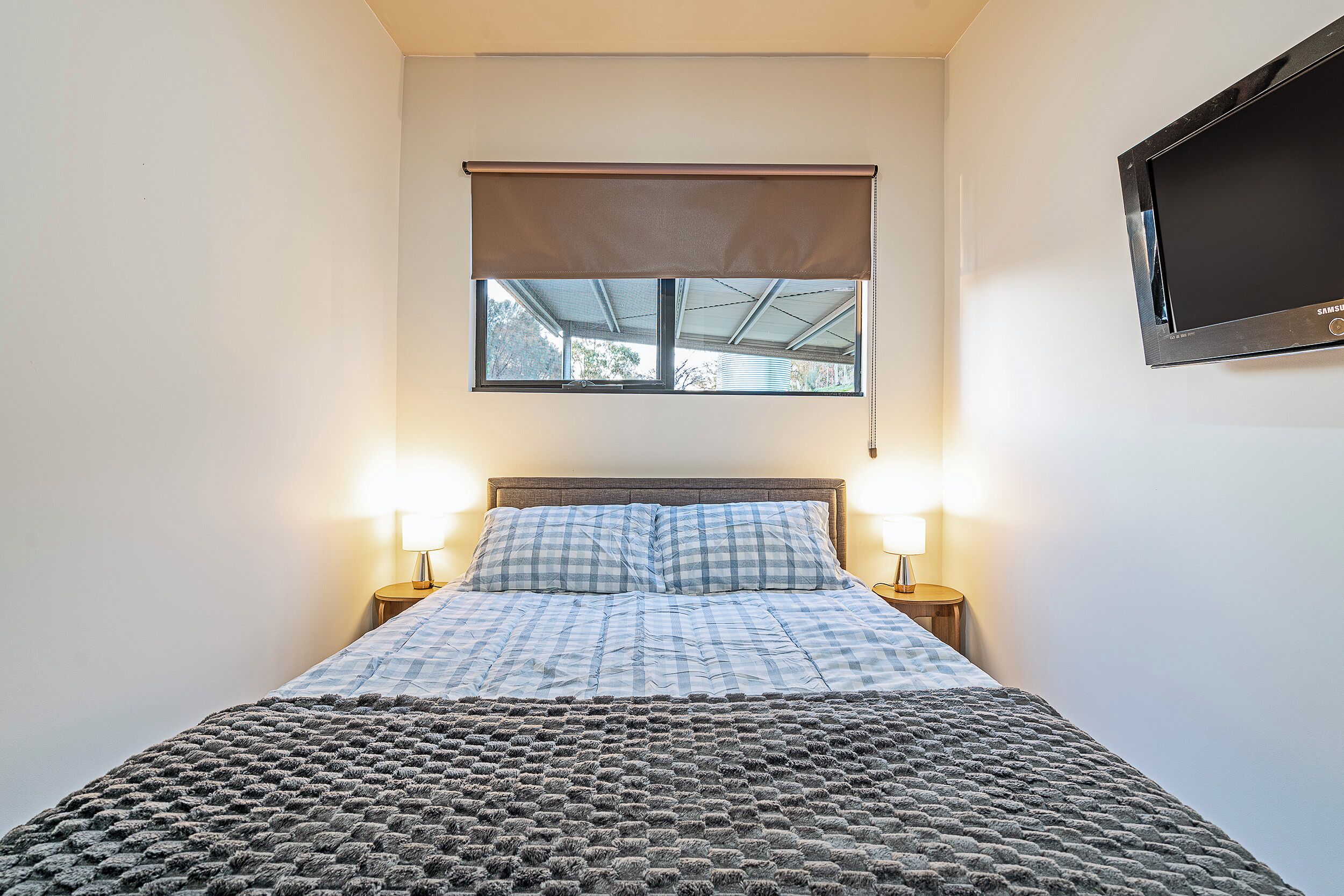 A bedroom with a bed, two bedside lamps, a window with a blind, and a television on the wall.