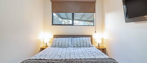 Une chambre à coucher avec un lit, deux lampes de chevet, une fenêtre avec un store et un téléviseur fixé au mur.