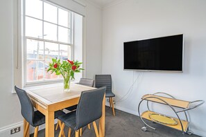 Una habitación con una mesa de madera, dos sillas, un florero con flores, un televisor de pantalla plana montado en la pared y una ventana con vista a edificios.
