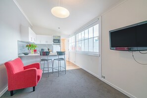 Una cocina moderna con armarios blancos, microondas y tetera. Hay una zona de comedor con una silla roja y una alfombra gris. Un ventanal grande proporciona luz natural.