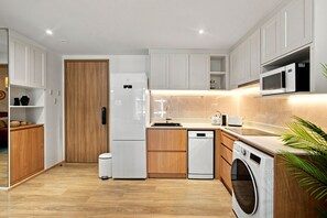 A modern kitchen with white cabinetry, a wooden door, a built-in dishwasher, a microwave, and a washing machine. - NEW! Apartment at Laguna Beachside: 2 bedrooms (Tambon Choeng Thale)