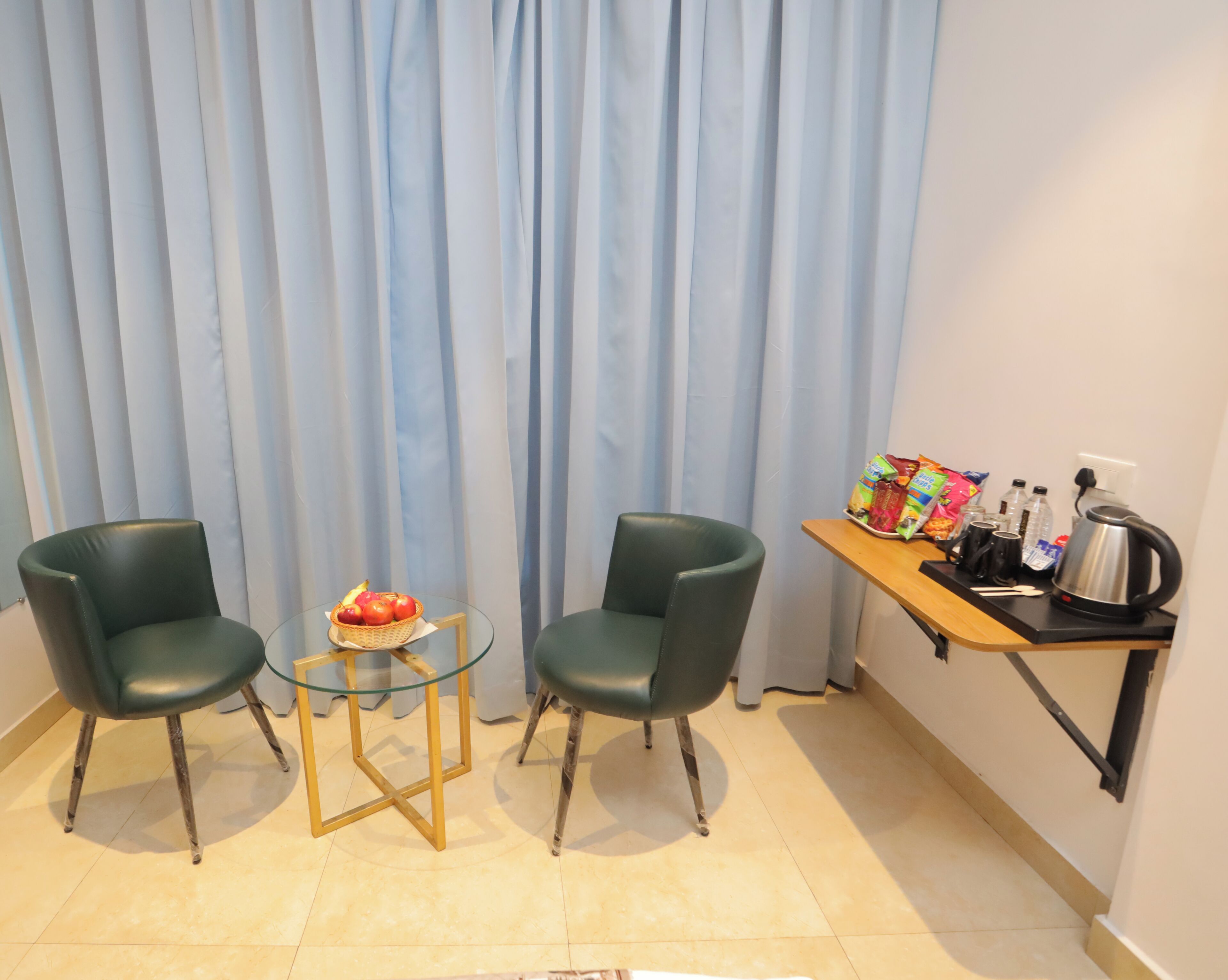 Een kleine kamer met twee stoelen, een glazen tafel, een fruitmand en een kitchenette met een waterkoker en lekkernijen.