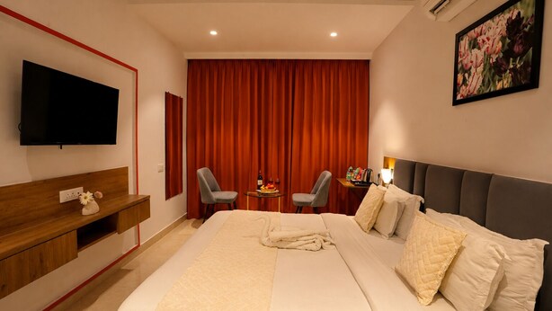 A hotel room with a large bed, a flat-screen TV, a desk, two chairs, a small table with a bottle and fruit, and a painting on the wall. - Hotel JHD Luxury Stay Restro & Sky Bar (Udaipur)
