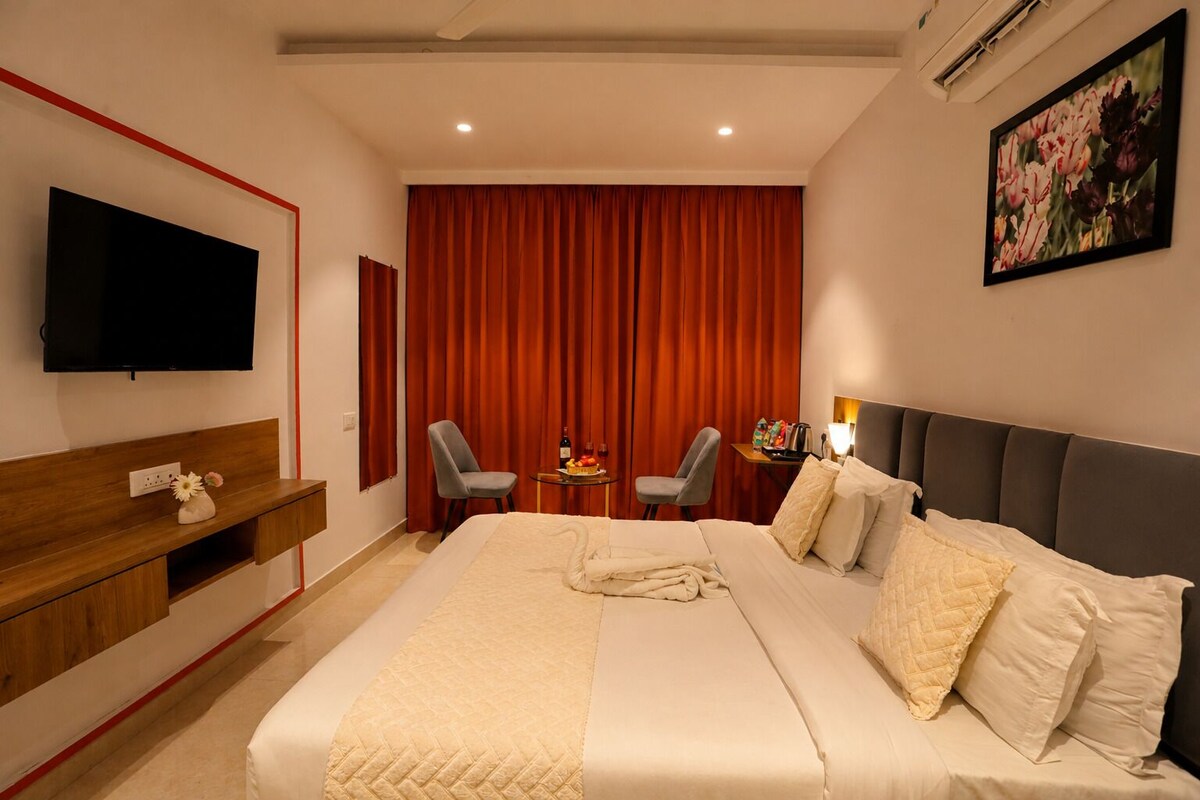 Ein Hotelzimmer mit einem großen Bett, einem Flachbildfernseher, einem Schreibtisch, zwei Stühlen, einem kleinen Tisch mit einer Flasche und Früchten sowie einem Bild an der Wand.