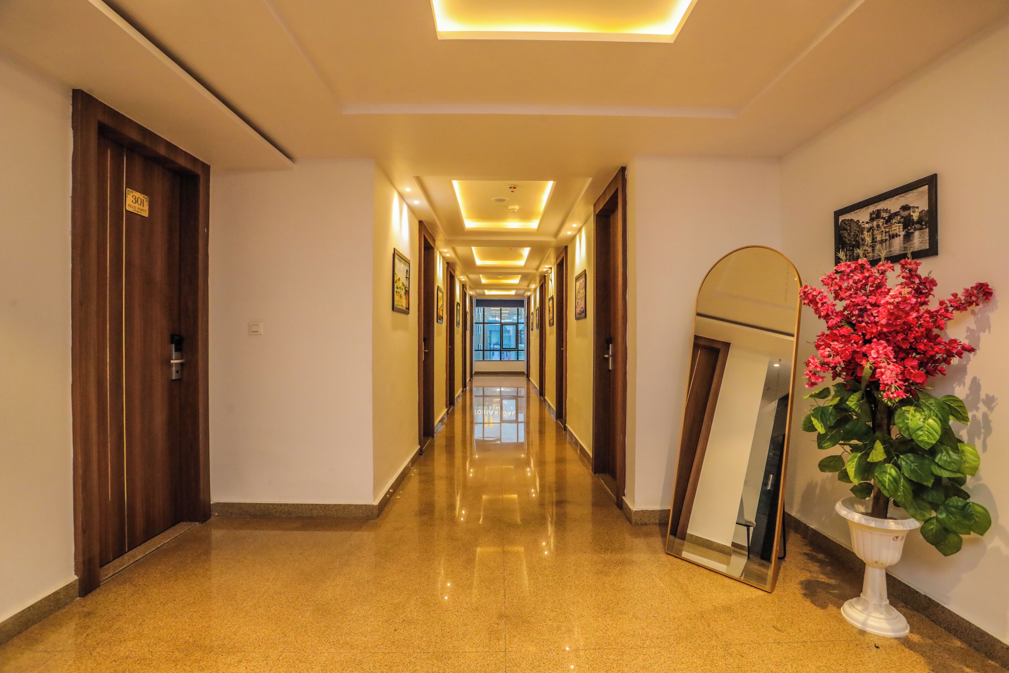 A hotel corridor with a polished floor, a wooden door on the left, a mirror on the right, and framed pictures on the walls.